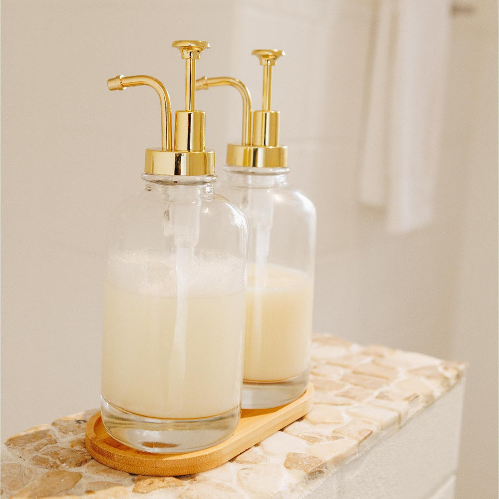 Clear glass dispenser bottle with gold pumps and bamboo tray